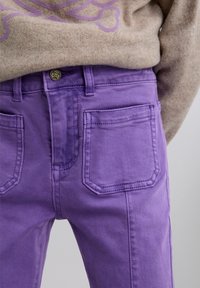Los pantalones vaqueros púrpura presentan una cintura alta, dos bolsillos traseros y un acento de botón con un diseño de calavera. Tienen una textura suave y un corte recto.