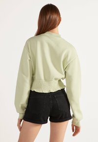 Světle zelená zkrácená mikina s nabíraným páskem a dlouhými rukávy, doplněná černými denimovými šortkami. Hladká textura, ležérní design.