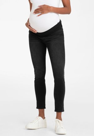 Gravid person iført sorte graviditetsjeans og hvide sneakers, der holder sin mave med begge hænder mod en ensfarvet baggrund.