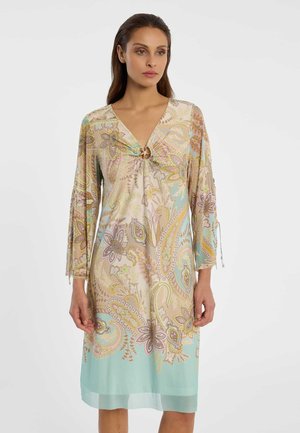 Frau trägt ein knielanges, langärmliges Kleid mit einem pastellfarbenen Paisley-Muster und einem Ringdetail am V-Ausschnitt.