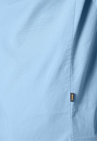 Ljusblått tyg med en slät yta, med en sydd söm och en liten svart och orange etikett på sidan.