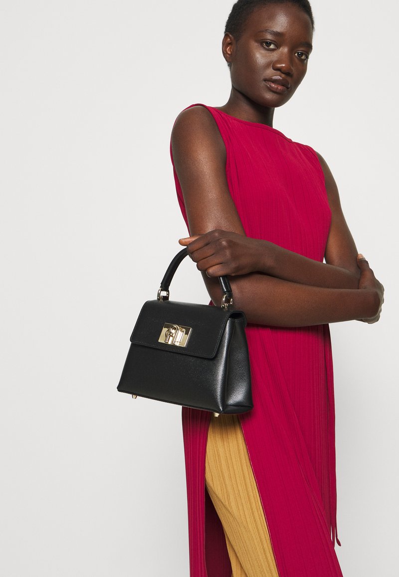 Bolsa de mão em couro preto com um design estruturado, ferragens douradas e uma alça superior. Vestido pregueado em vermelho e amarelo complementa a bolsa.