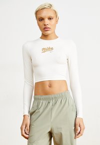Ilgas baltas crop topas su aukso ir rudos spalvos Nike logotipu ant krūtinės, derinamas su laisvais, šviesiai žaliais sportiniais džinsais, turinčiais baltą logotipą.