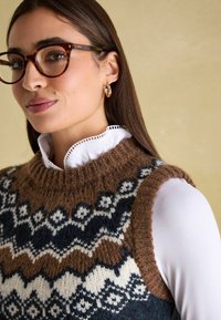 Braune, gestrickte Weste mit einem geometrischen Muster in Marineblau und Weiß, ausgestattet mit einem hohen, gerippten Kragen und ärmellosem Design über einer weißen Bluse.