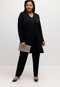 Túnica negra de mangas largas con escote en V y detalle de botones, combinada con pantalones negros y tacones negros. Un bolso de mano gris se sostiene en una mano.
