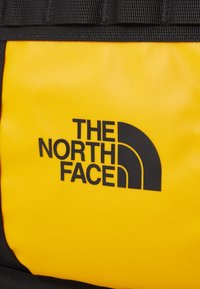 Gul rygsæk med et sort logo med tre buede linjer og teksten: "THE NORTH FACE." Holdbart stof med en glat tekstur.