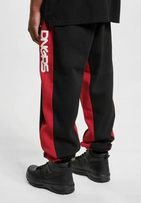 Black sweatpants with red side stripes featuring a white logo. Material appears cotton blend. Black boots with laces are worn.
