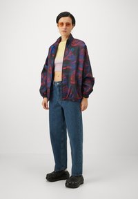 Printed bomber jacket in dark fabric with red and blue patterns. Paired with light yellow cropped top, high-waisted jeans, and black clogs.