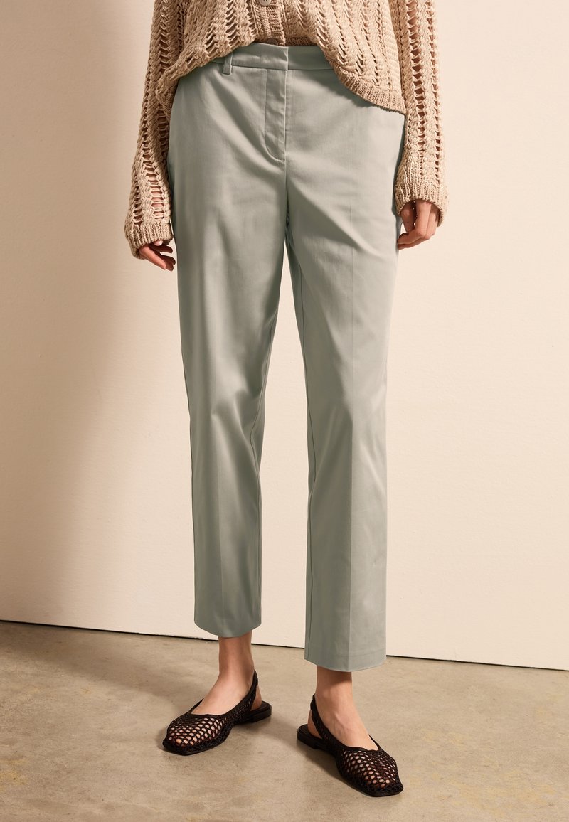 Hellgrüne, maßgeschneiderte Hose mit glatter Textur und geradem Schnitt, kombiniert mit schwarzen geflochtenen Ballerinas. Ein gestrickter beiger Pullover sorgt für Kontrast.