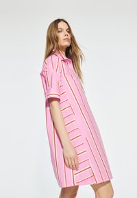 Růžové šaty košilového střihu se svislými a vodorovnými červenými a bílými pruhy. Krátké rukávy, límečkový design a uvolněný střih.