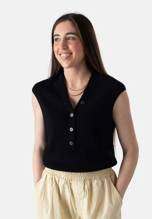 Joven mujer con cabello castaño largo sonriendo, vestida con una blusa negra sin mangas con botones, pantalones beige y collares de cadenas doradas, con las manos en los bolsillos.