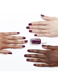 Diep paarse nagellak in een doorzichtige fles met een witte dop, omringd door twee paren handen met gemanicuurde diepe paarse nagels op een witte achtergrond.
