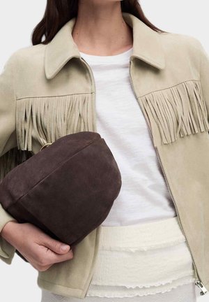 Vrouw draagt een beige jas met franjes en een wit topje, houdt een grote donkerbruine suède clutch tegen haar zij vast.