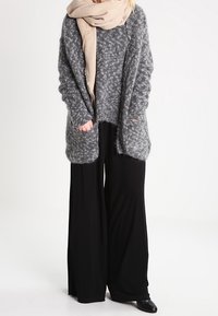 Šedý pletený cardigan s volným střihem, který má přední kapsy. Nositý přes černý top a široké černé kalhoty. Béžový šátek přehozený kolem krku.