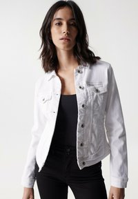 Chaqueta de mezclilla blanca con cuello, bolsillos frontales y herrajes plateados. Presenta un diseño ajustado y cierre de botones con mangas largas.