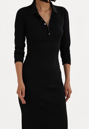 Femme portant une robe noire à manches longues avec col boutonné et silhouette ajustée, sur un fond blanc uni.