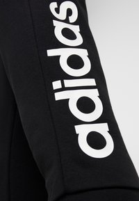 Camisa negra de manga larga con el logotipo blanco de "adidas" impreso verticalmente a lo largo de la manga izquierda. La textura de la tela parece suave y elástica.