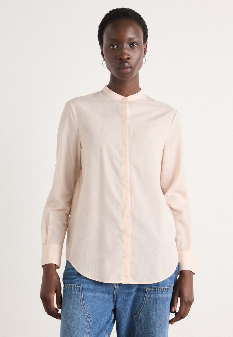 Camicia rosa chiaro a maniche lunghe con colletto alla mandarino, chiusura con bottoni e bottoni a contrasto, abbinata a jeans denim azzurri.