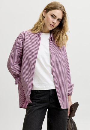 Junge Frau mit langen blonden Haaren, die ein lila gestreiftes Hemd über einem weißen T-Shirt und schwarzen Hosen trägt und eine braune Jacke in der Hand hält.