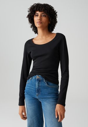 Vrouw met krullend haar draagt een nauwsluitend zwart shirt met lange mouwen en blauwe jeans met hoge taille tegen een effen grijze achtergrond.