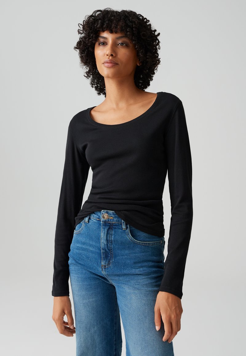 Vrouw met krullend haar draagt een nauwsluitend zwart shirt met lange mouwen en blauwe jeans met hoge taille tegen een effen grijze achtergrond.