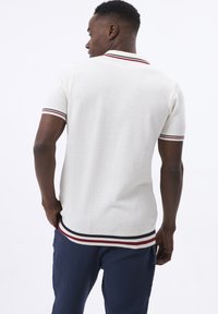 Camiseta polo blanca con un tejido texturizado, que presenta un cuello y puños de manga acanalados con rayas rojas y marinas. Llevada con pantalones marinos.