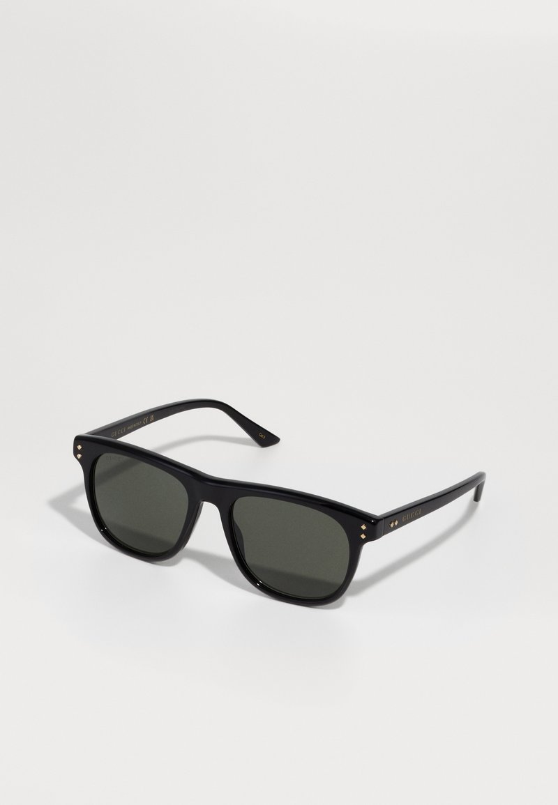 Ochelari de soare din plastic negru, cu lentile verde închis, rame pătrate și accente aurii pe sprijinul umerilor. Textură netedă și design minimalist.