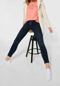 Mörkblå skinny jeans med hög midja och femknäppt stängning, kombinerat med en korallfärgad t-shirt och vita sneakers, sittande på en svart pall.