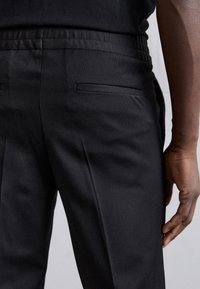 Pantalon noir en tissu texturé, avec une taille élastique et une poche arrière unique. La coupe est ajustée slim.