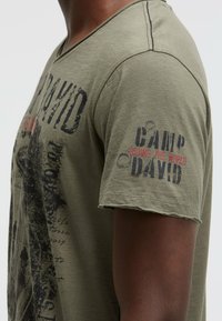 Olive green short-sleeve T-shirt made of soft cotton, featuring a V-neck, distressed printing of "CAMP DAVID," and subtle stitching detail.