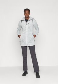 Giacca con cappuccio foderata in pile di colore grigio chiaro, con zip frontale, due tasche laterali e coulisse regolabili. Abbinata a pantaloni scuri e stivali neri.