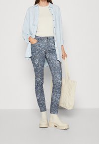 Ljusblå skjorta över en krämfärgad topp, kombinerad med mönstrade blå jeans och beige klumpiga stövlar. Beige toteväska i handen.