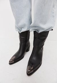 Bottes de cowboy en cuir noir à bouts pointus, avec un embout métallique et un talon bloc, portées avec un jean en denim bleu clair décontracté.