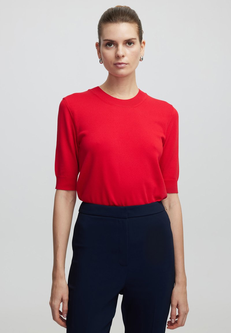 Suéter corto de manga corta en rojo, hecho de una tela de punto suave, con un cuello redondo y un corte relajado, combinado con pantalones oscuros de color marino.