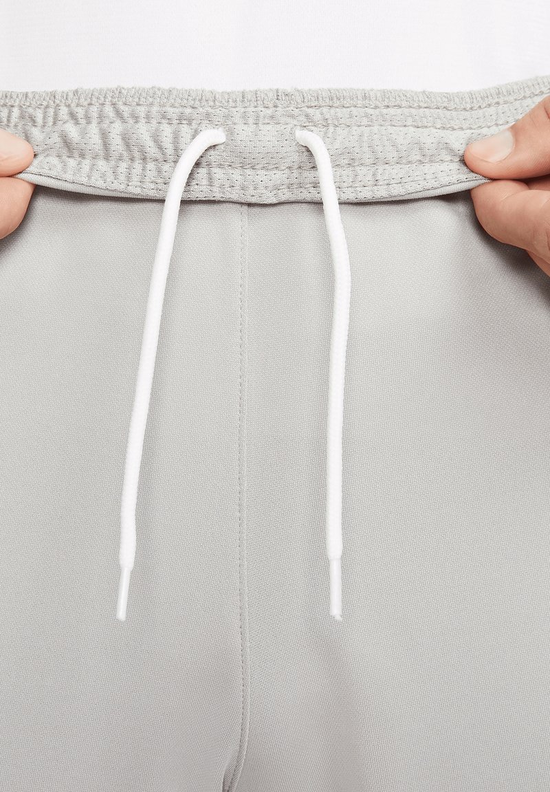 Mains tenant la ceinture élastique grise d’un pantalon de jogging avec des cordons blancs centrés au-dessus de la couture.