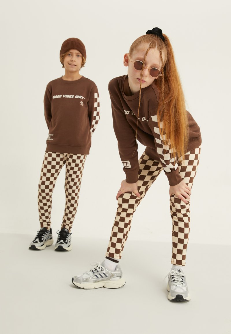 Rjava pulover s tekstom "Samo dobre vibracije", v kombinaciji z rjavimi in smetano karirastimi leggings. Obe imata športne superge, ena pa nosi kapo.