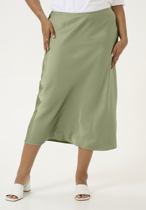 Woman wearing olive green midi skirt, white top, and white open-toe heeled mules, standing with hands on hips.