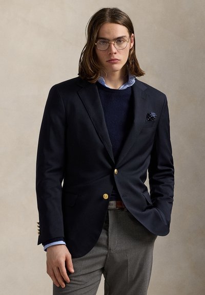 Ung mand med skulderlangt hår iført runde briller, en navy blazer med guldknapper, lyseblå skjorte, navy sweater og grå bukser.