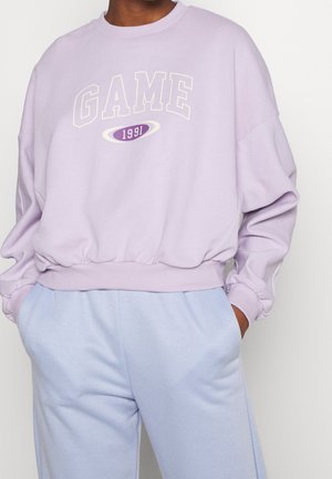 Person wearing light purple sweatshirt with "GAME 1991" text and light blue pants, hands in pockets, against plain background.