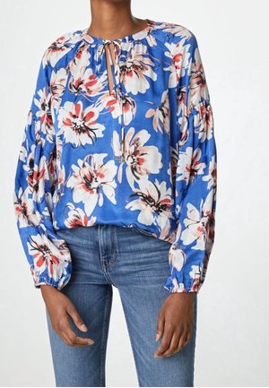 Blusa azul con gran estampado floral en blanco y rojo, mangas largas abullonadas y escote en forma de ojo de cerradura, combinada con vaqueros azules.