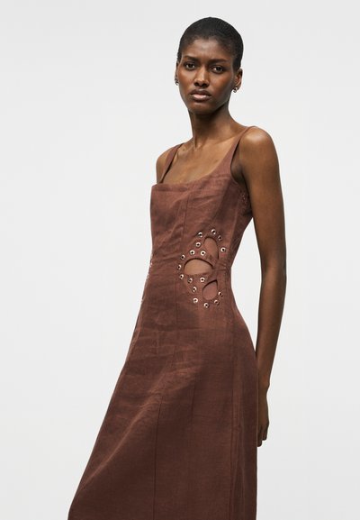 Γυναίκα που φοράει καφέ αμάνικο φόρεμα με ανοίγματα και μεταλλικά κορδόνια στη μέση, στέκεται μπροστά σε απλό γκρι φόντο.