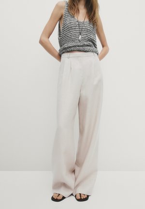 Vrouw draagt een grijze gebreide mouwloze top, beige wijde pijpen broek, zwarte sandalen en een hangende ketting, staand tegen een effen achtergrond.