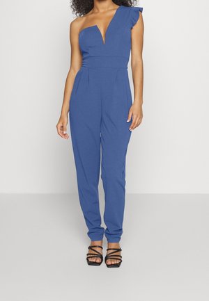 Donna che indossa una tuta blu monospalla con scollatura profonda e sandali neri con tacco e cinturini, in piedi davanti a uno sfondo semplice.