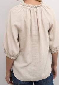 Blouse en lin beige avec un col froncé, des manches bouffantes trois-quarts et des poignets élastiques. Texture douce et ourlet légèrement incurvé à l'arrière.