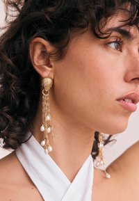 Gold-tone earrings with a scalloped design and hanging chains, adorned with small white pearls. The overall look is flowing and textured.