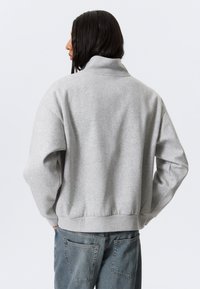 Persoon met lang donker haar, gekleed in een lichtgrijze trui met hoge kraag en blauwe spijkerbroek, die met de rug naar de kijker staat en met zijn handen in de zakken.