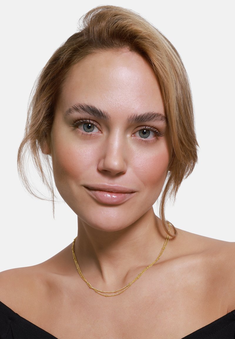 Collana d'oro a due strati con catene sottili e una superficie liscia, indossata su una clavicola scoperta, su uno sfondo bianco.