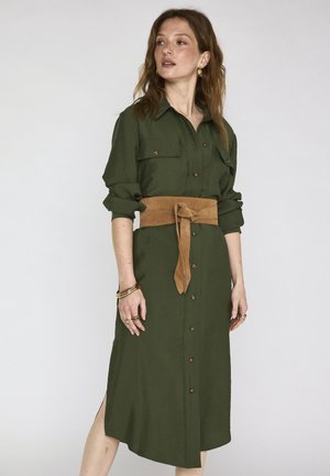 Donna che indossa un vestito camicia color verde oliva con maniche arrotolate e una ampia cintura in camoscio color tan annodata in vita, accessoriata con braccialetti e anelli dorati.