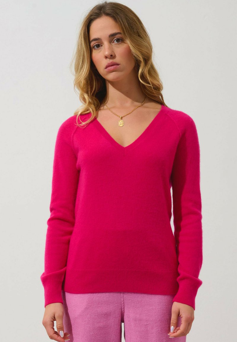 Pull V en fuchsia en tricot doux avec des manches longues ; poignets et ourlet côtelés ; porté avec un pantalon rose clair ; texture lisse.