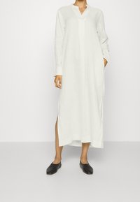 Robe longue en lin blanc avec un col à revers, des boutons, des fentes latérales et des manches longues. Associée à des chaussures noires sans lacets.